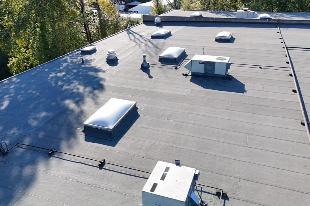 Flat commercial building roof with several HVAC units, vents, and skylights; surrounded by trees and neighboring structures, photographed in daylight from an elevated angle.