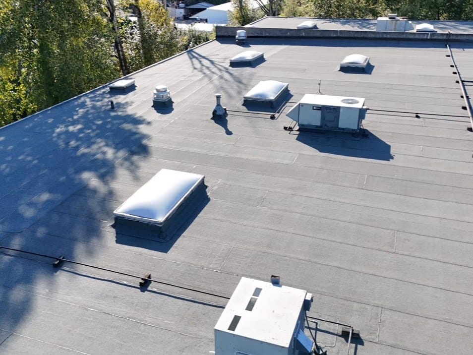 Flat commercial building roof with several HVAC units, vents, and skylights; surrounded by trees and neighboring structures, photographed in daylight from an elevated angle.