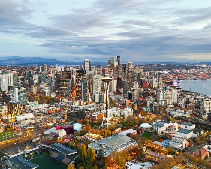 Aerial view of downtown Seattle showing the city skyline with the Space Needle, tall buildings, and waterfront under a cloudy sky, with city lights starting to glow at dusk.