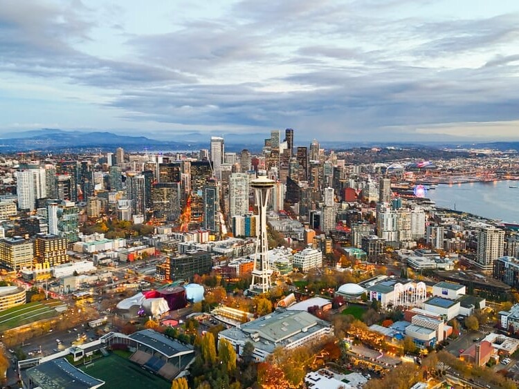 Aerial view of downtown Seattle showing the city skyline with the Space Needle, tall buildings, and waterfront under a cloudy sky, with city lights starting to glow at dusk.