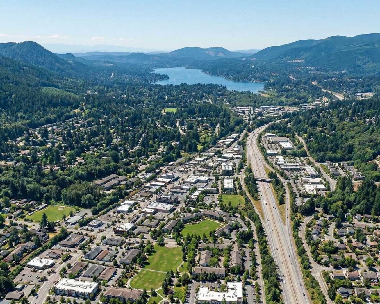 Aerial view of a suburban area with a main road running through the center, surrounded by houses, green trees, and hills, with a large lake visible in the background.