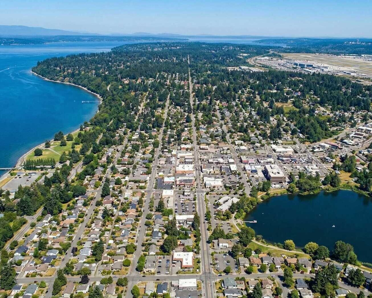 Aerial view of a suburban coastal city with a grid of houses, a large body of water on the left, a small lake on the right, and an airport in the distance under a clear blue sky.