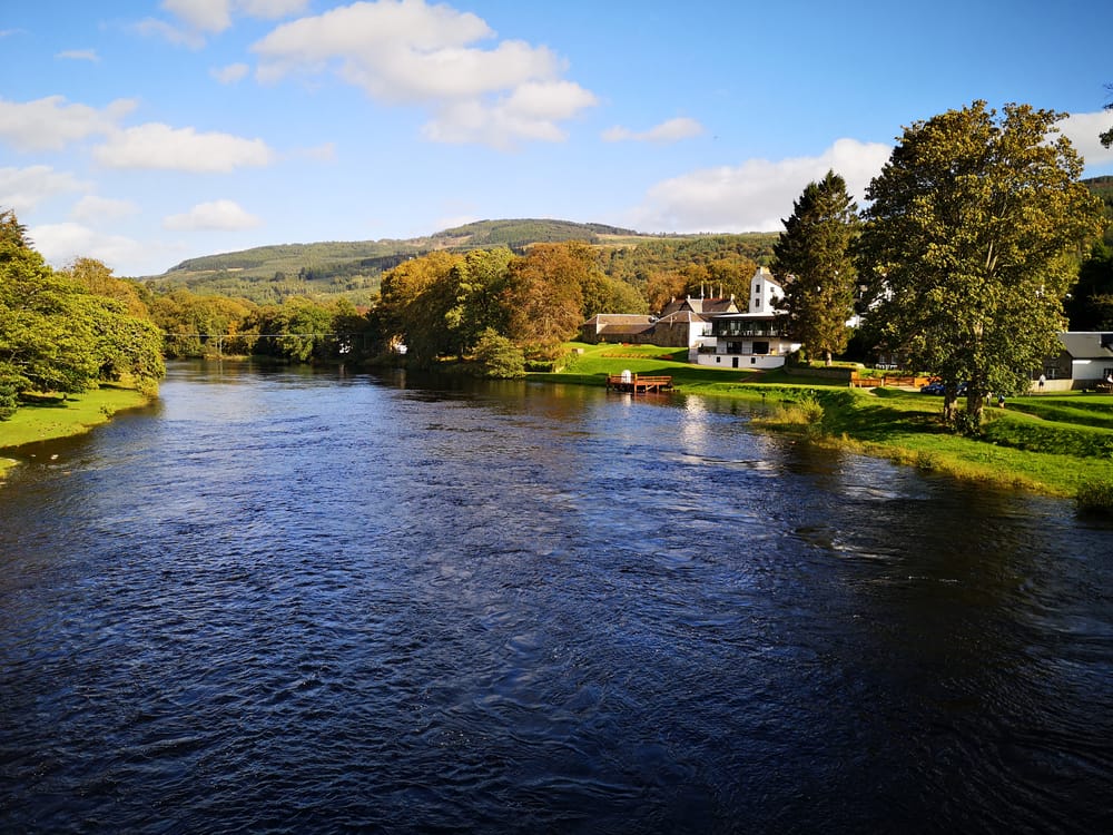 A wide river flows past green trees and grassy banks, with white houses and a small dock on the right. Hilltops covered in trees rise in the background under a blue sky with fluffy clouds.