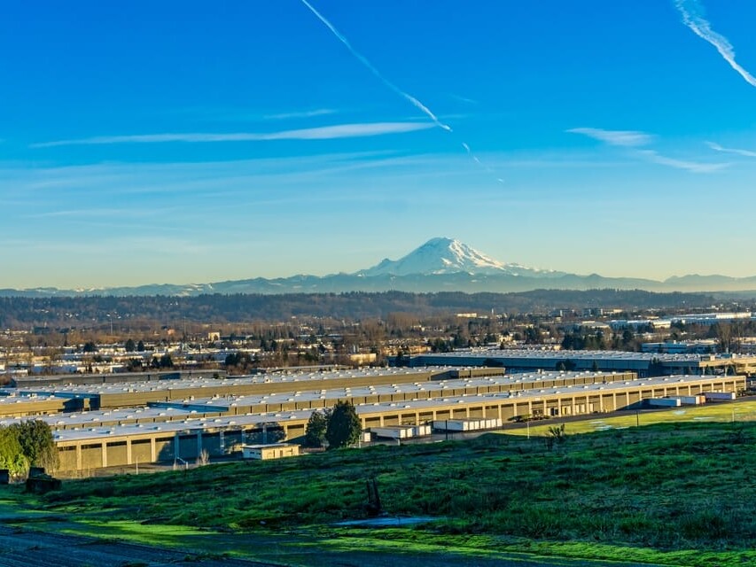A wide view of an industrial complex with green grass in the foreground and a snow-capped mountain in the background under a clear blue sky with visible contrails.