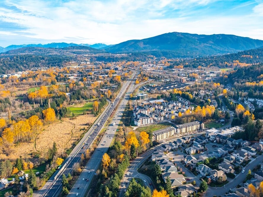 Aerial view of a suburban neighborhood with houses, tree-lined streets, and a highway running through the center, surrounded by autumn foliage and mountains in the distance under a blue sky.