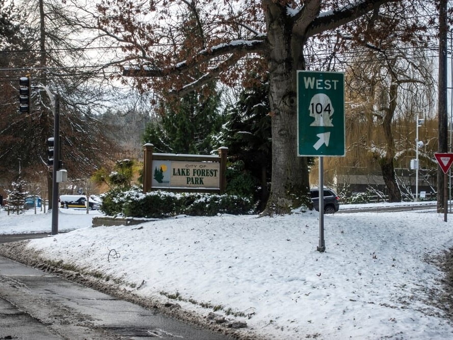 A snowy street scene shows a City of Lake Forest Park sign near a large tree, with a green West 104 highway sign and a yield sign. Snow covers the ground and trees, with traffic lights and cars in the background.